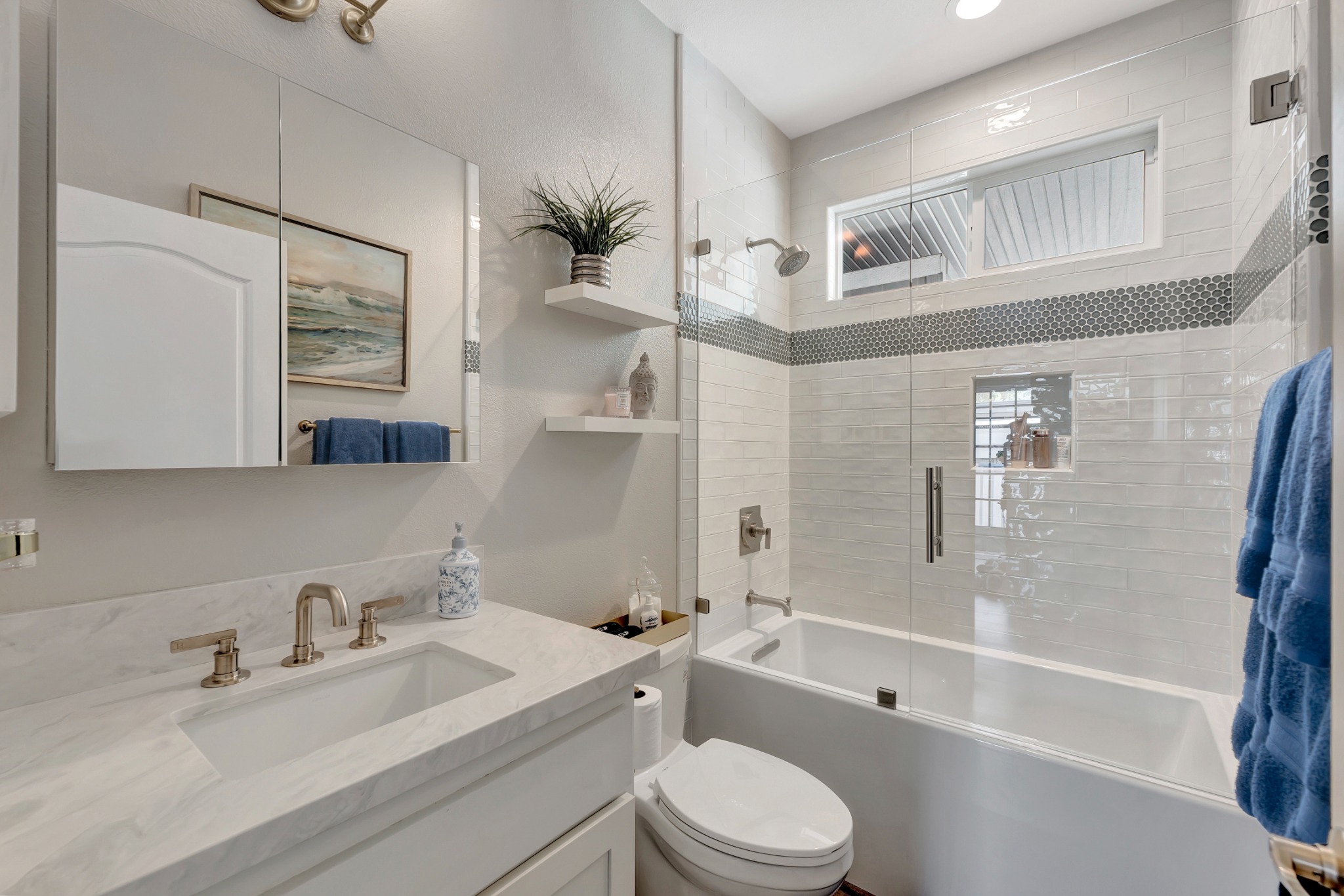 Bright Newport Beach guest bathroom remodel featuring white vanity with marble-style countertop, brushed nickel faucet, floating shelves, and subway tile shower with mosaic accent band.