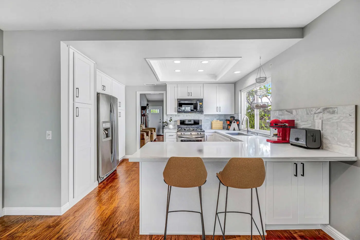 A bright, modern kitchen remodel in Irvine with white cabinetry, quartz countertops, natural wood barstools at the peninsula, and hardwood floors filled with natural light.