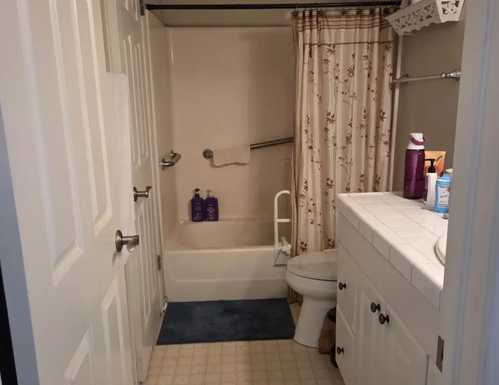 Before photo of an Irvine bathroom with an older tub-shower combo, white tiled vanity, floral shower curtain, and dated flooring.