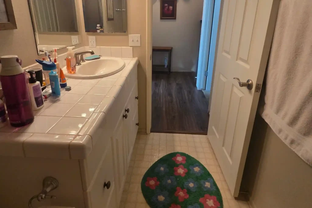 Before photo of an Irvine bathroom with a white vanity, tiled countertop, older mirror, beige flooring, and dated fixtures.