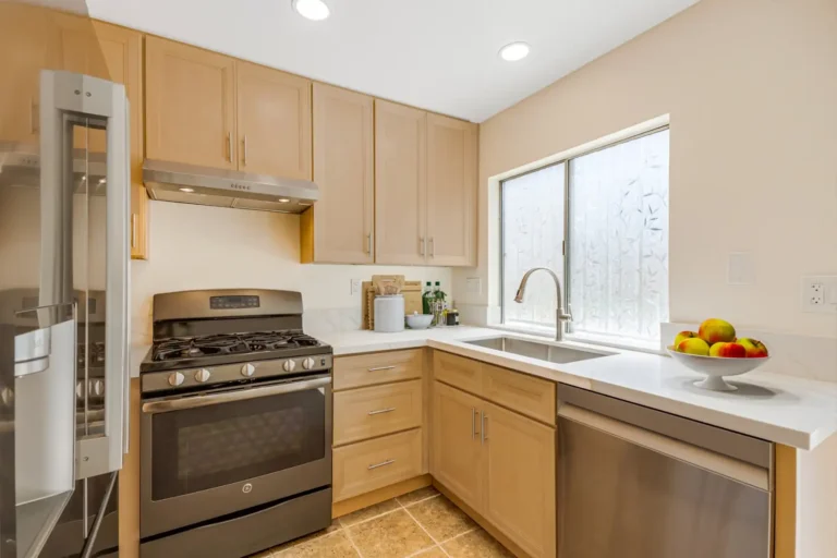 Close-up of an Irvine kitchen sink with natural wood cabinets, white quartz countertops, stainless steel dishwasher, and window above.