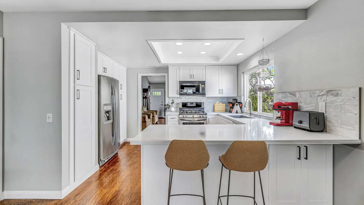 White shaker cabinets and a quartz peninsula with two counter stools in an Irvine kitchen remodel with wood floors.