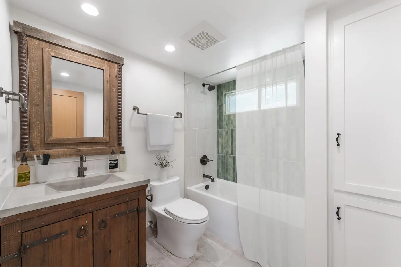 Rustic wood vanity with a framed mirror beside a green accent tile tub shower in a remodeled Santa Ana guest bathroom.