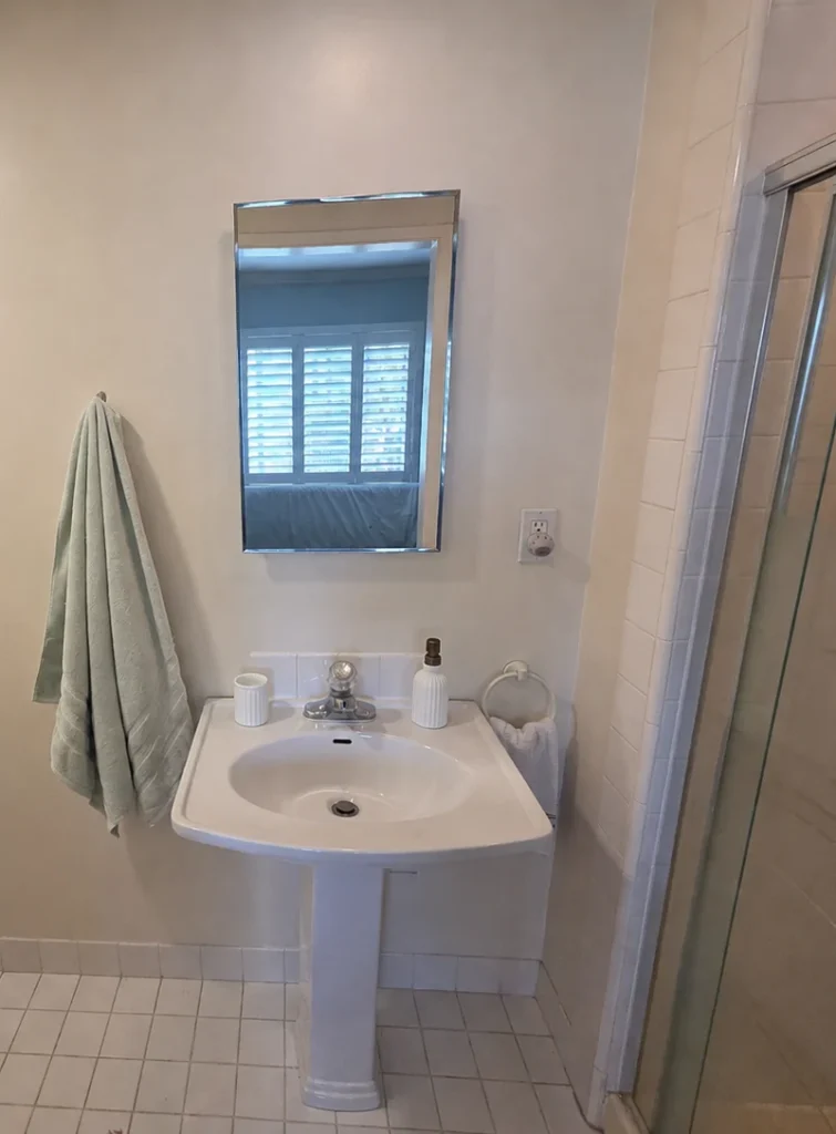 Before photo of a Cypress master bathroom showing the original pedestal sink, rectangular mirror, white tile floor, and framed shower enclosure.