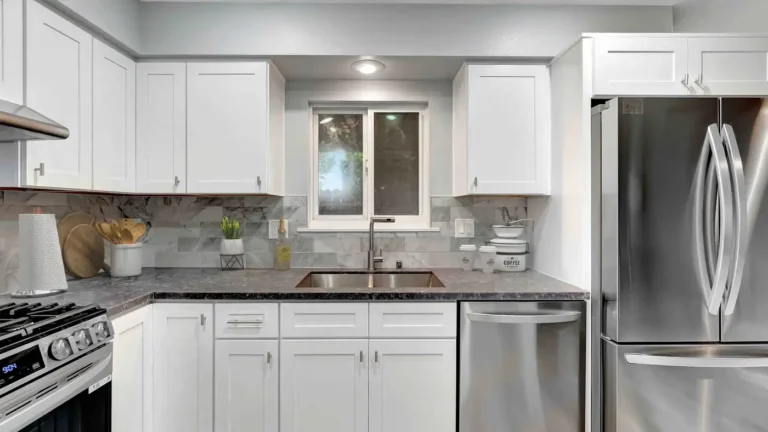 Double stainless sink under a window with marble-look backsplash and white shaker cabinets in a Huntington Beach kitchen