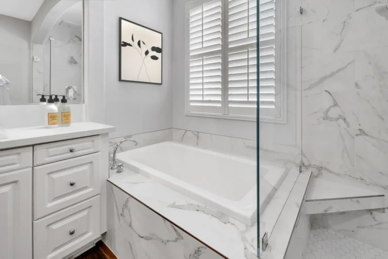 Drop-in soaking tub with a marble-look tile deck below plantation shutters in a Carlsbad primary bathroom.