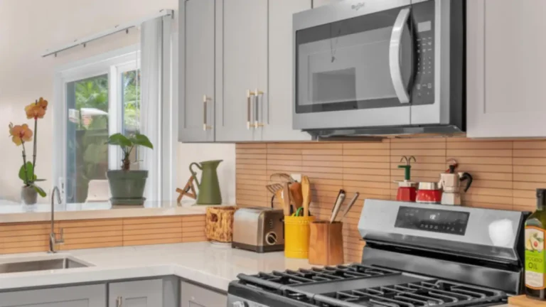 Warm tan stacked tile backsplash behind a gas range and microwave with gray shaker cabinets in a Carlsbad kitchen