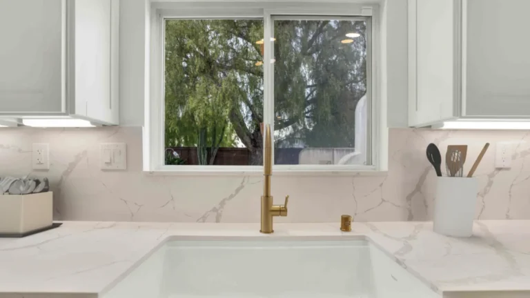 Brushed brass faucet over a white farmhouse sink with a quartz slab backsplash in an Oceanside kitchen remodel