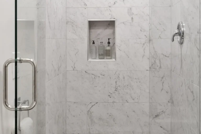 Marble tile shower wall with a recessed shampoo niche, chrome valve and glass door in an Irvine bathroom remodel
