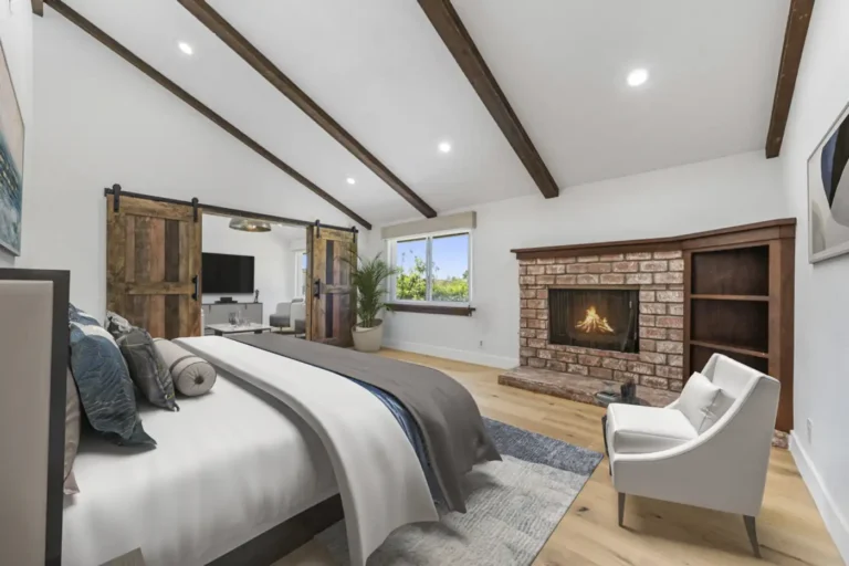 Santa Ana primary bedroom with dark wood ceiling beams, brick fireplace, rustic barn doors, and wide-plank wood floors.