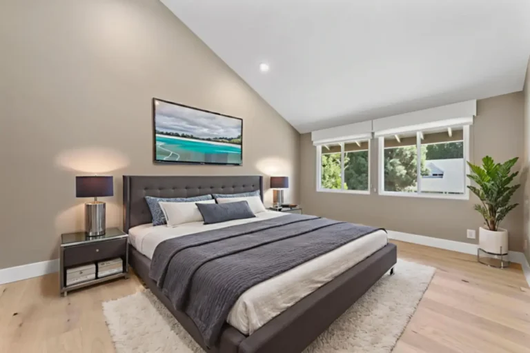 Santa Ana bedroom with taupe walls, a vaulted ceiling, tufted gray bed, light wood flooring, and two white windows.